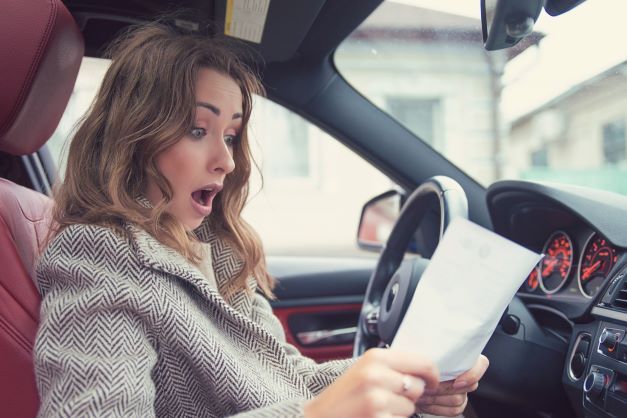 Woman sitting in driver's seat of car, shocked at the paperwork she is reviewing.
